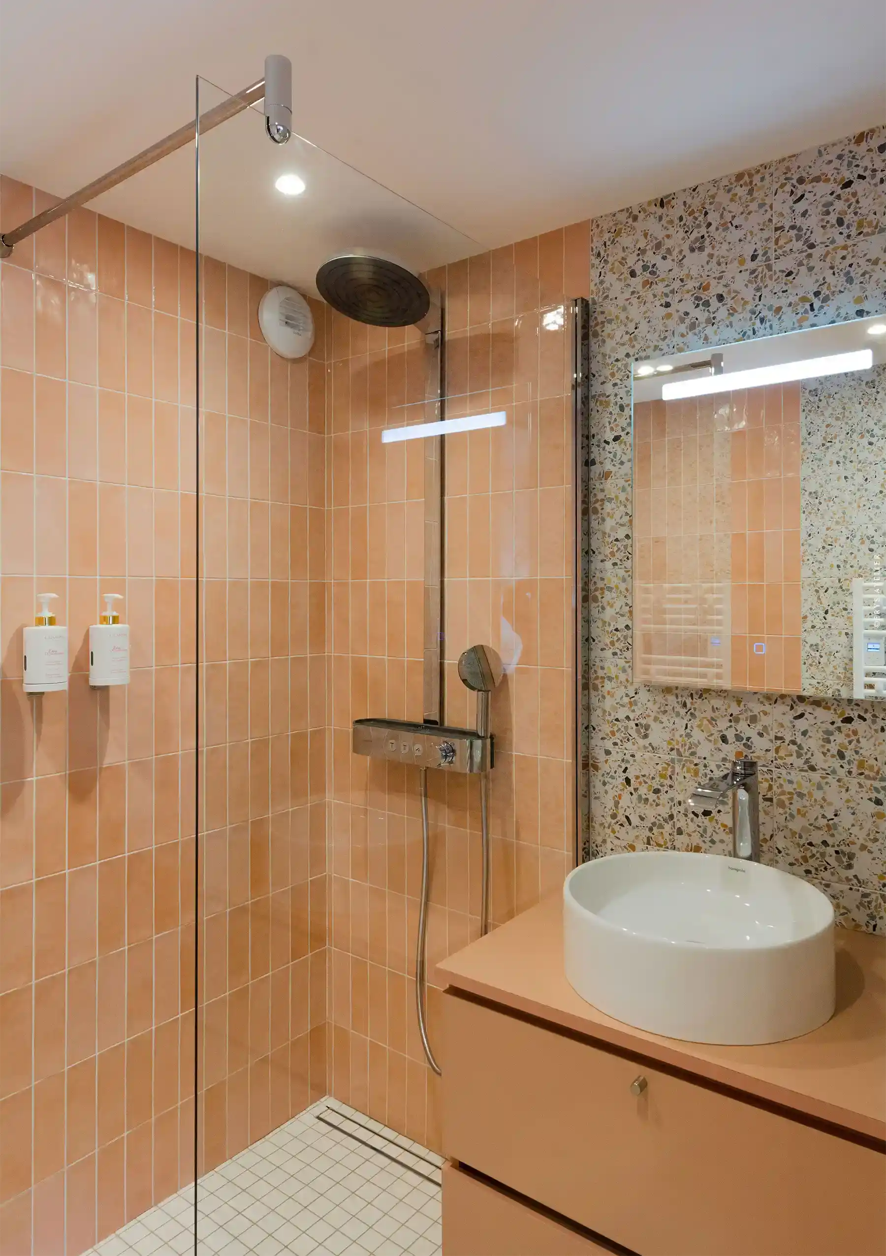 Lavabo rond blanc et douche à l'italienne avec murs en carreaux peach et mur en terrazzo. Salle de bain moderne avec éclairage intégré.