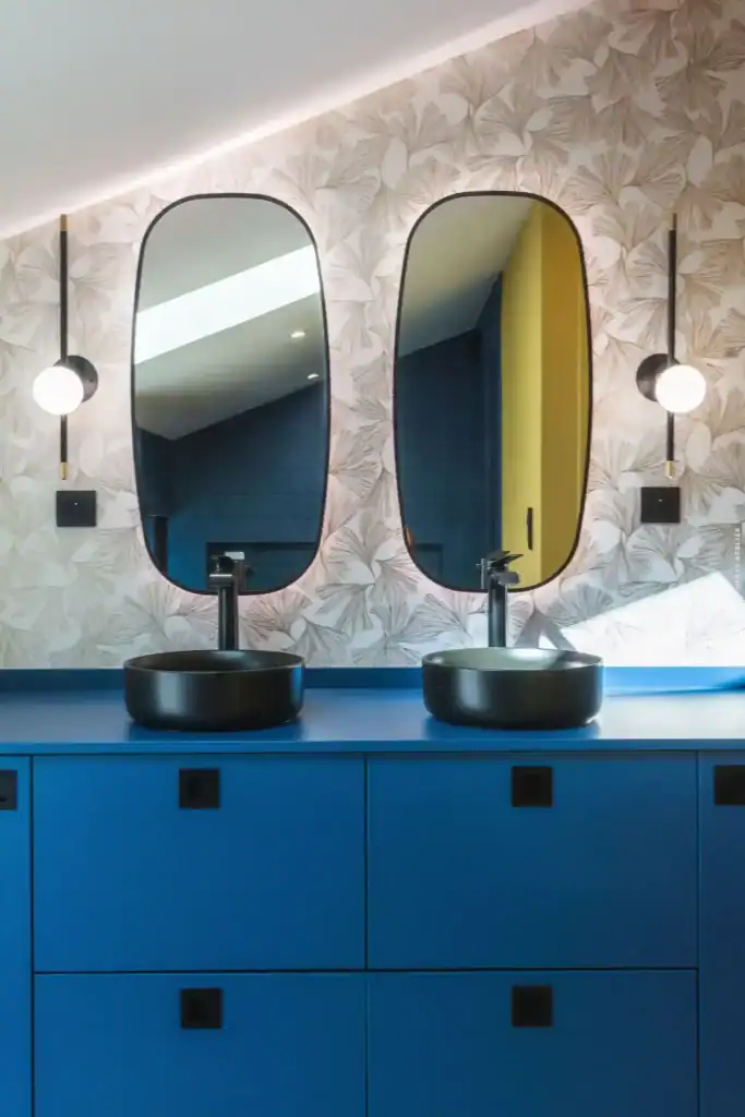 Baigneuses noires avec lavabos sur meuble bleu, murs avec papier peint à motifs végétaux et miroirs ovales.