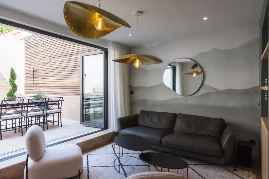 Moderne intérieur avec canapé en cuir gris et décoration design, vue sur terrasse avec mobilier extérieur, mur peint avec paysage abstrait, luminosité chaleureuse.
